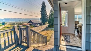 A wooden deck with a view of a road and distant buildings, a door leading to an interior with a lamp and a chair. - Near Snoqualmie River! Cozy Home in Rural Setting (Carnation)