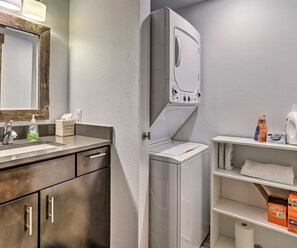 A bathroom with a wooden vanity, a sink, a mirror, a built-in washing machine, and shelves with toiletries. - Main Street Frisco Condo w/ Private Patio! (Frisco)