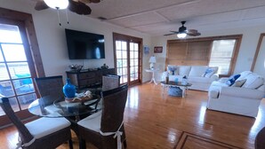 A spacious living room with a wooden floor, a dining area with wicker chairs, a white sofa, and a flat-screen TV mounted on the wall. - Mermaid House: Heated Pool, Dual Dockage & Outdoor Bar on the Canal (Ramrod Key)