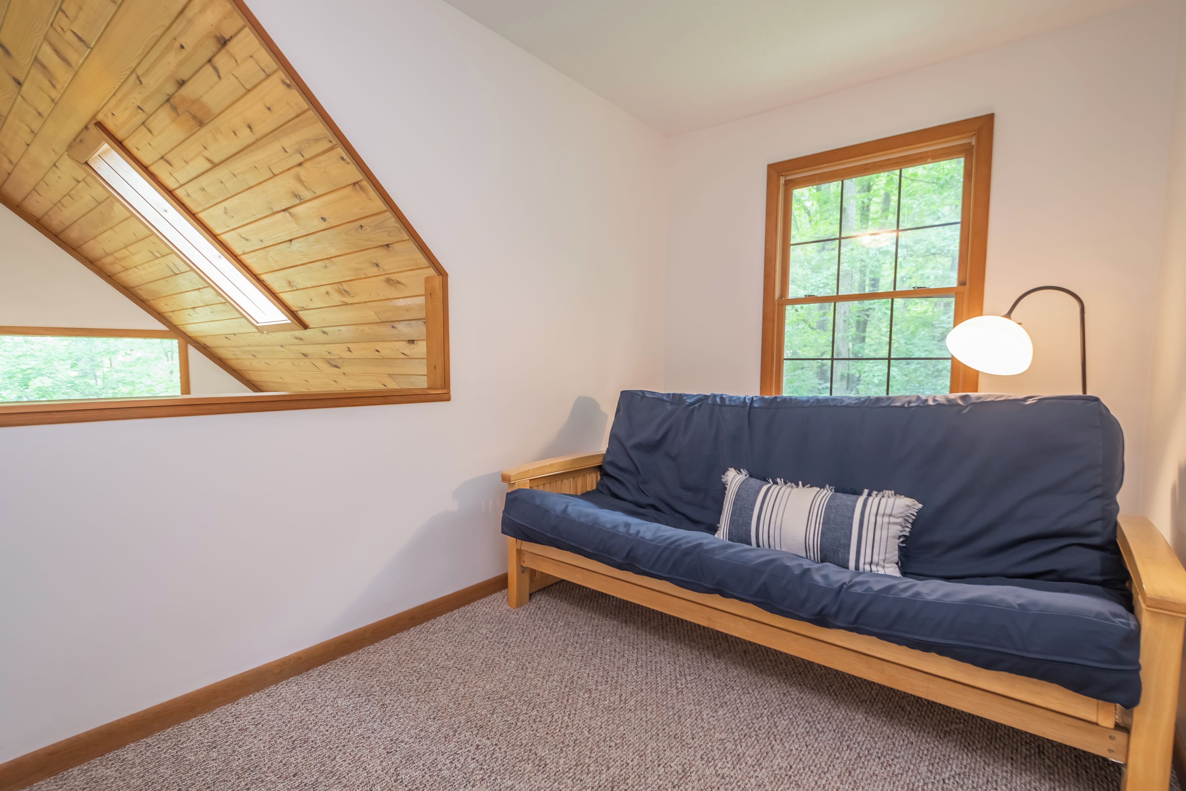 A room with a wooden sloped ceiling, a window with a view of trees, a dark blue sofa with a lamp, and a carpeted floor.