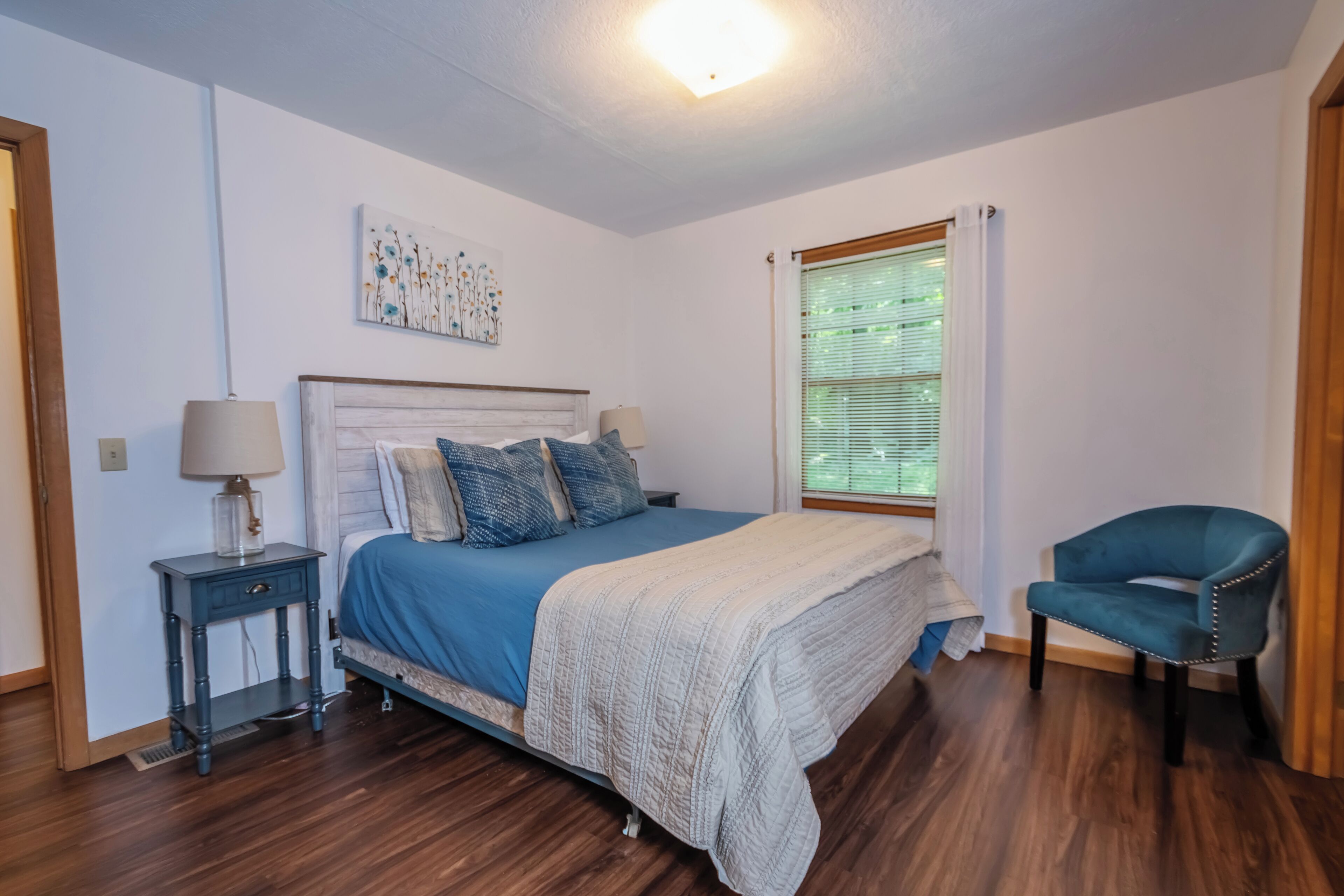 A bedroom with a bed, a nightstand, a chair, a window with blinds, and a ceiling light.