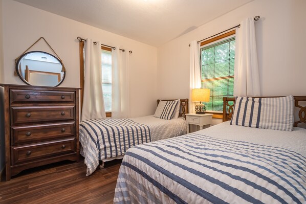 A bedroom with two beds, a wooden dresser, a mirror, a nightstand with a lamp, and a window with blinds.