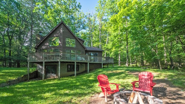 Uma cabana com varanda, cercada por árvores, e duas cadeiras Adirondack vermelhas perto de um braseiro.