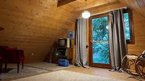 Ein gemütliches Blockhausszimmer mit Holzwänden und einer geneigten Decke. Es verfügt über ein großes Fenster mit Vorhängen, einen Fernseher, einen kleinen Tisch und einen Schaukelstuhl.