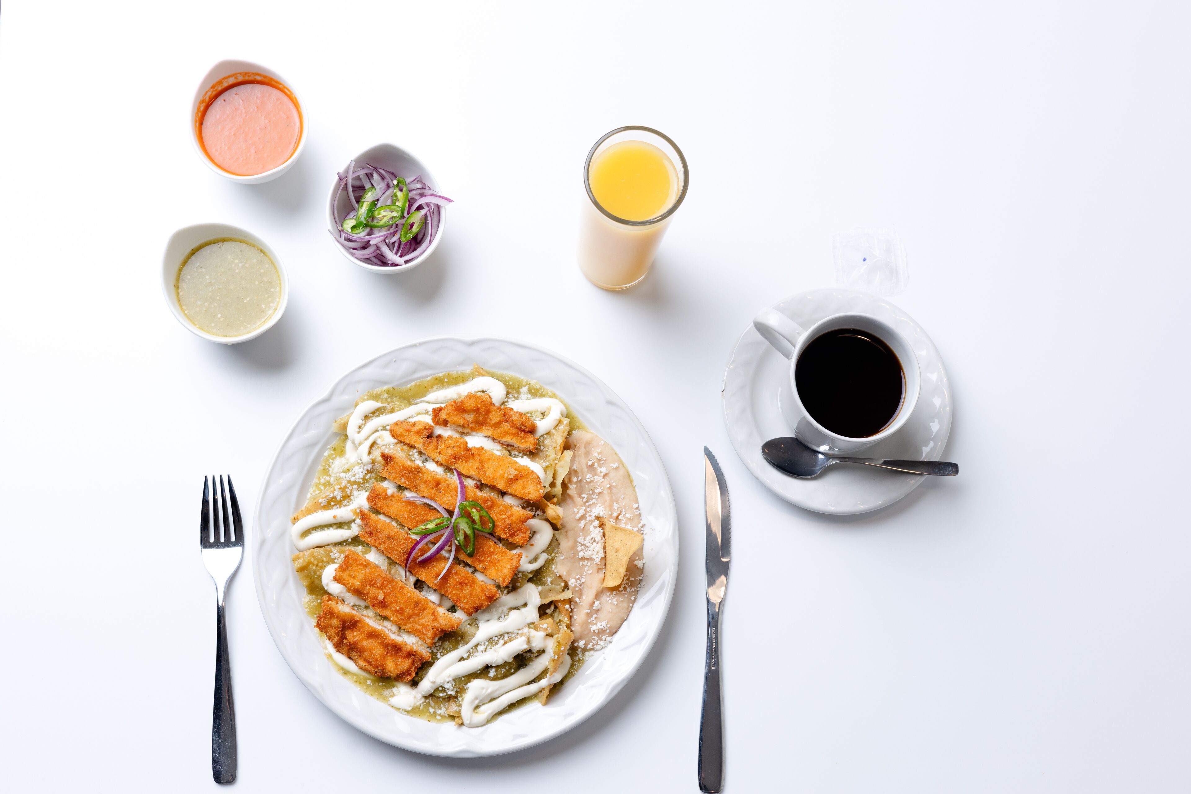A plate of food with breaded cutlets, a glass of orange juice, a cup of black coffee, and small bowls of condiments.