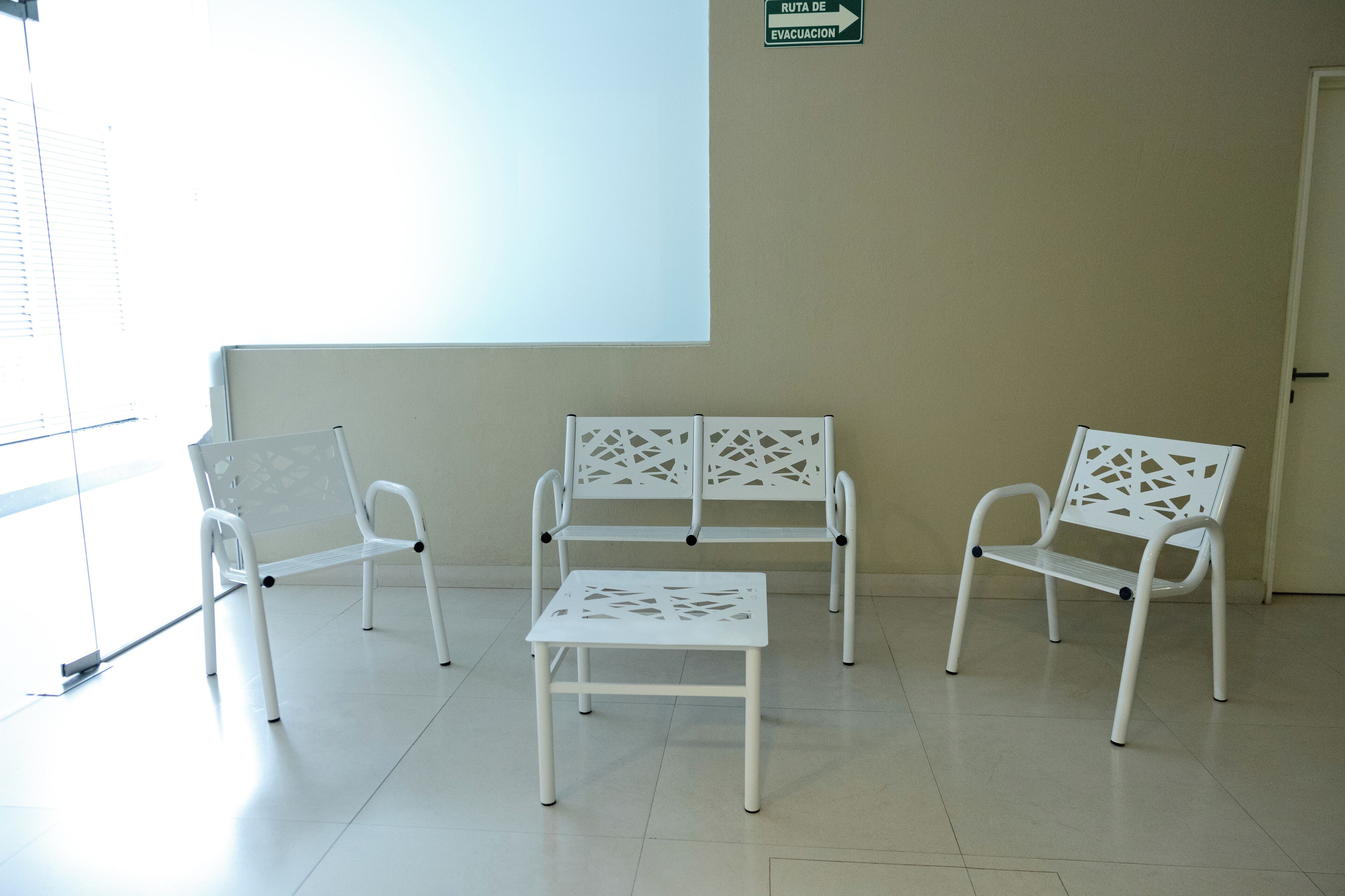 Uma área de espera com cadeiras brancas, uma mesa, tela de projeção e sinalização de rota de evacuação.