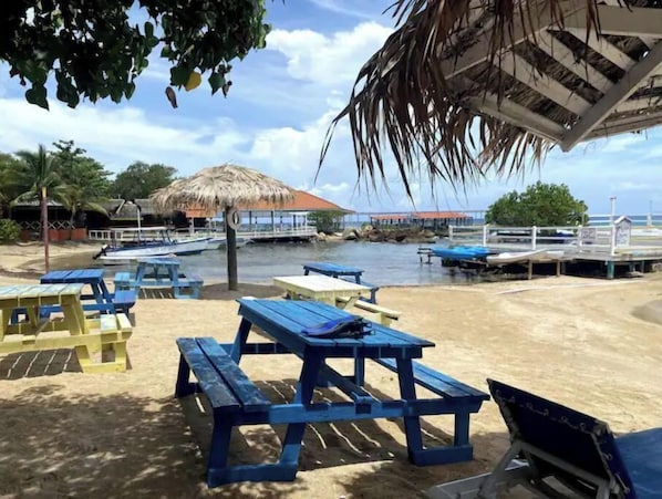 A beach with blue picnic tables, a thatched-roof structure, and boats in the background.