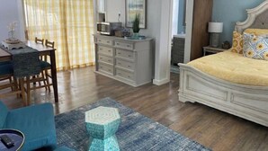 A bedroom with a bed, bedside table, lamp, and a dresser. There is a dining area with a table and chairs.