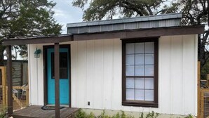 A small, single-story building with a white exterior and a blue door. It features a wooden deck and a stone foundation.