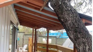 A covered outdoor area with a wooden bench, a tree, and a view of a blue house in the background.