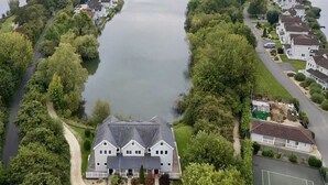 A large house with a dark roof, surrounded by lush greenery and a calm lake. - Spring Lake 77, The Ferns (P) - Lakeside Luxury, Hot Tub, Watersports, Sleeps 8 (South Cerney)