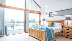 A bedroom with a wooden bed, a nightstand with lamps, and a large window overlooking a body of water. - Waters Edge 05, Amaranth Lodge (P) - Lakeside Holiday Home, Hot Tub, Sleeps 6 (Cirencester)