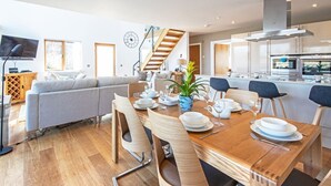 A modern living room with a sofa, a dining table set with plates and glasses, a TV, and a staircase leading to another level. - Waters Edge 05, Amaranth Lodge (P) - Lakeside Holiday Home, Hot Tub, Sleeps 6 (Cirencester)