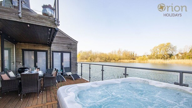 A wooden deck with a hot tub and outdoor seating, overlooking a body of water. - Waters Edge 05, Amaranth Lodge (P) - Lakeside Holiday Home, Hot Tub, Sleeps 6 (Cirencester)