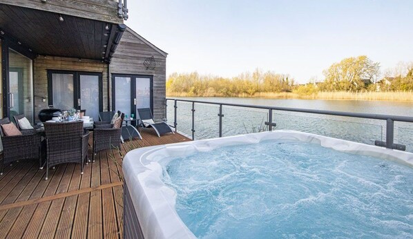 Terraza de madera con jacuzzi y mobiliario exterior, con vistas a un cuerpo de agua.