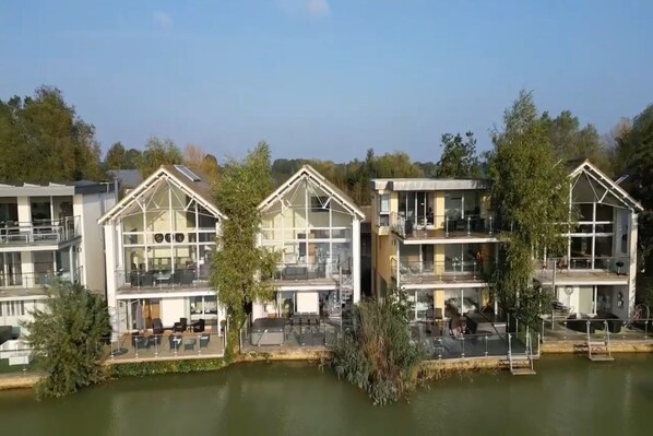 A row of modern, glass-fronted buildings with balconies, situated by a calm body of water, likely a lake or river. - Howells Mere 26, Swans Down - Lakeside House, Hot Tub, Spa Access, Sleep 8-10 (Somerford Keynes)