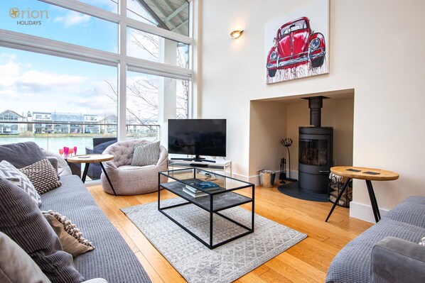A modern living room with a large TV, comfortable seating, a fireplace, and a view of a body of water. - Howells Mere 26, Swans Down - Lakeside House, Hot Tub, Spa Access, Sleep 8-10 (Somerford Keynes)