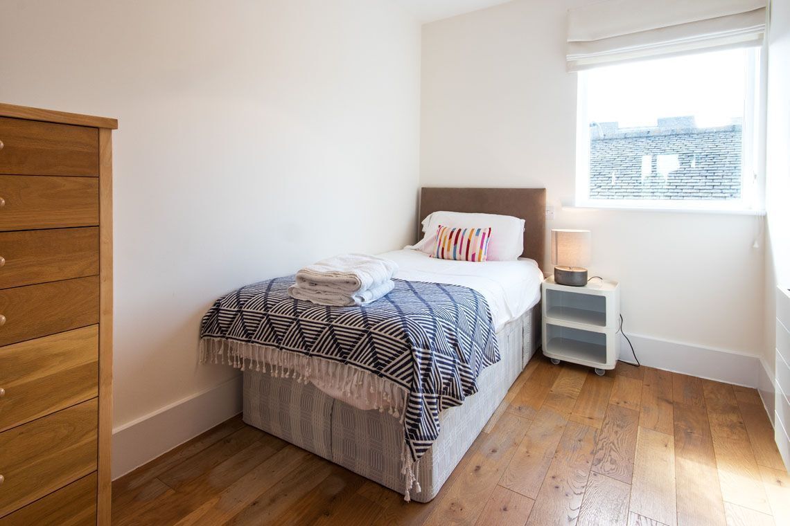 Un dormitorio con cama, cómoda de madera, mesita de noche con lámpara y ventana con vista a edificios.