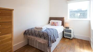 Un dormitorio con cama, cómoda de madera, mesita de noche con lámpara y ventana con vista a edificios.