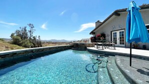 A clear swimming pool with a stone edge, a patio with a table and chairs, and a blue umbrella. - House with pool/spa/minutes to wineries (Temecula)