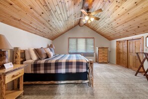 Une chambre avec un plafond en bois, un lit recouvert d’une couverture à carreaux, une table de chevet, une commode et une fenêtre avec des stores.