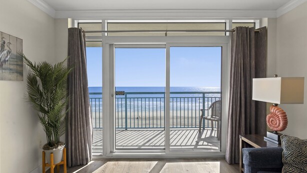 Ein Zimmer mit einer großen Schiebeglasdoor zu einem Balkon mit Blick auf einen Strand. Auf dem Balkon befinden sich ein Blumentopf, eine Couch und eine Lampe.