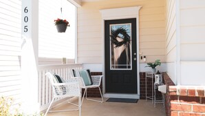 A front porch with a black door, white wicker chairs, a potted plant, and a brick wall.