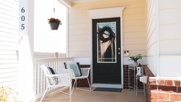 A front porch with a black door, white wicker chairs, a potted plant, and a brick wall.