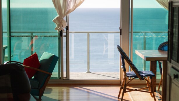 A room with a large window overlooking the ocean, a chair, a table, and a view of the sea.