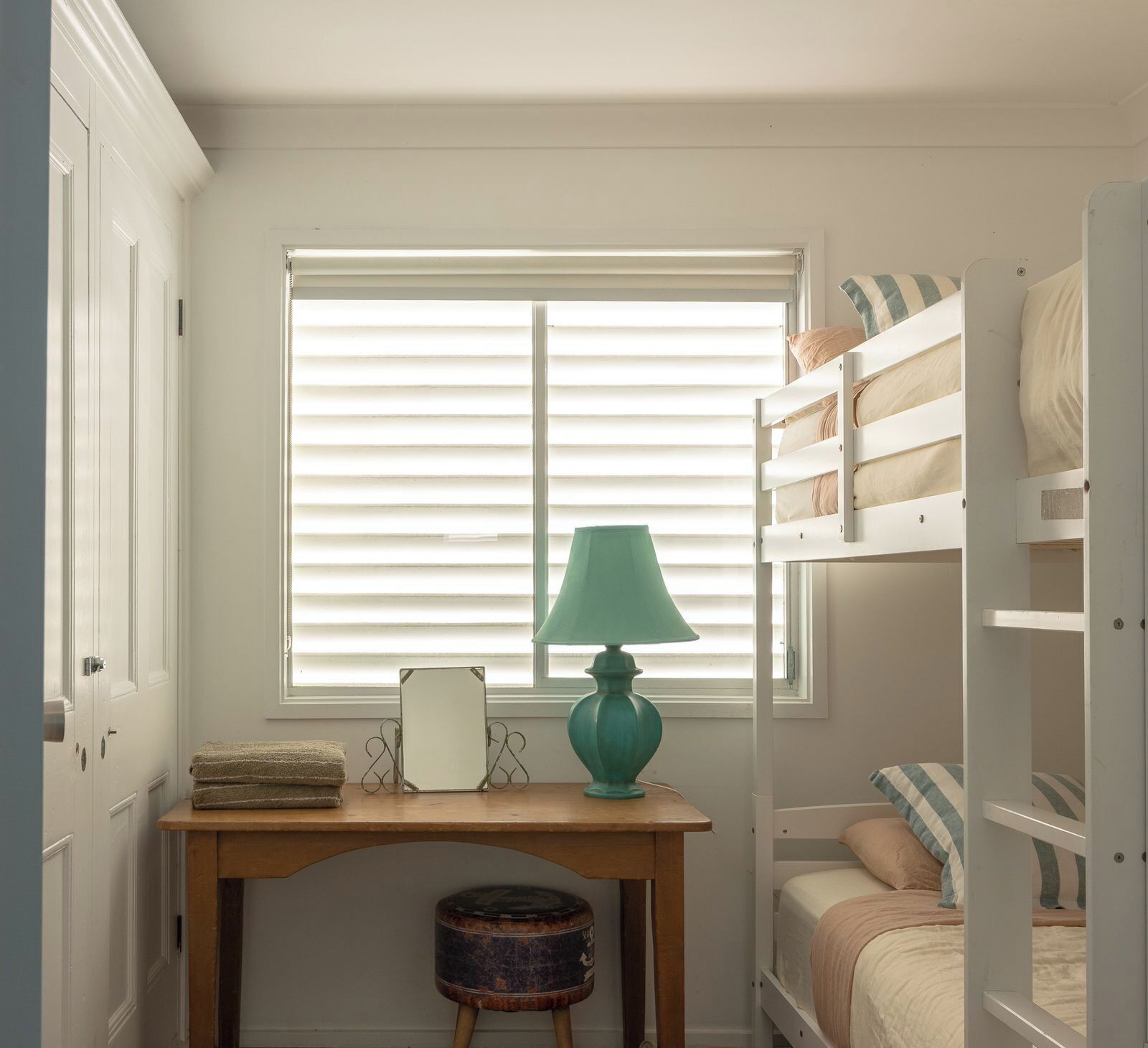 A room with a window, a desk with a lamp, a small stool, and a bunk bed with pillows.
