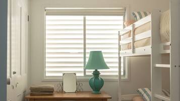 A room with a window, a desk with a lamp, a small stool, and a bunk bed with pillows.
