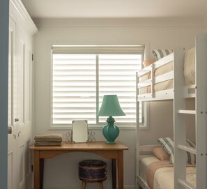 Une pièce avec une fenêtre, un bureau avec une lampe, un petit tabouret et un lit superposé avec des oreillers.