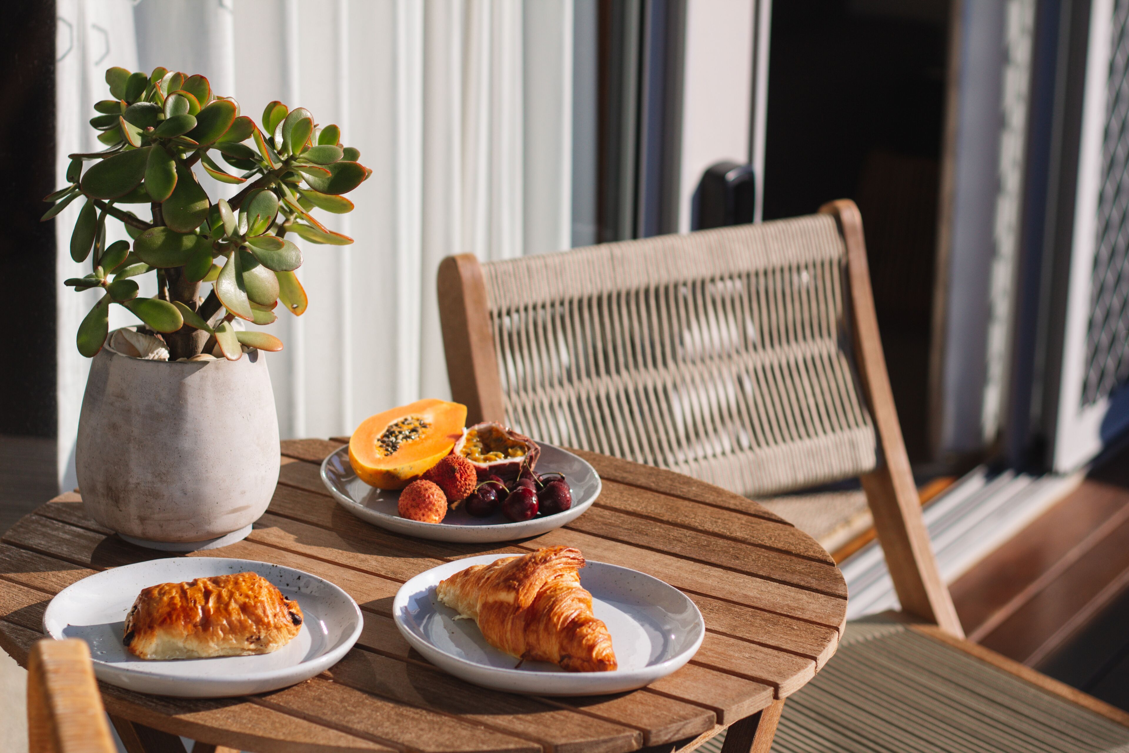 A round wooden table with two plates of food, one with a croissant and the other with fruit and a pastry. A potted plant is also visible.