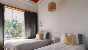 A bedroom with two beds, a large window with curtains, a white brick wall, and a hanging light fixture. - BAY PROPERTIES | The Loft Yamba (Yamba)