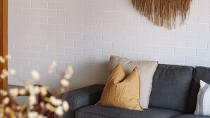A grey sofa with two cushions against a white brick wall, a woven wall hanging above, and a blurred plant in the foreground. - BAY PROPERTIES | The Loft Yamba (Yamba)