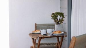 A wooden table with croissants and white cups, a wicker chair, and a potted plant on a balcony with white walls. - BAY PROPERTIES | The Loft Yamba (Yamba)