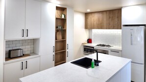 A modern kitchen with white cabinets, a wooden shelf, a built-in oven, and a stainless steel refrigerator. - U Suites on Jessie (Wellington)