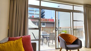 A modern room with a sliding glass door leading to an outdoor balcony. Inside, there is a sofa with colorful cushions, a chair, and a table. - U Suites on Jessie (Wellington)
