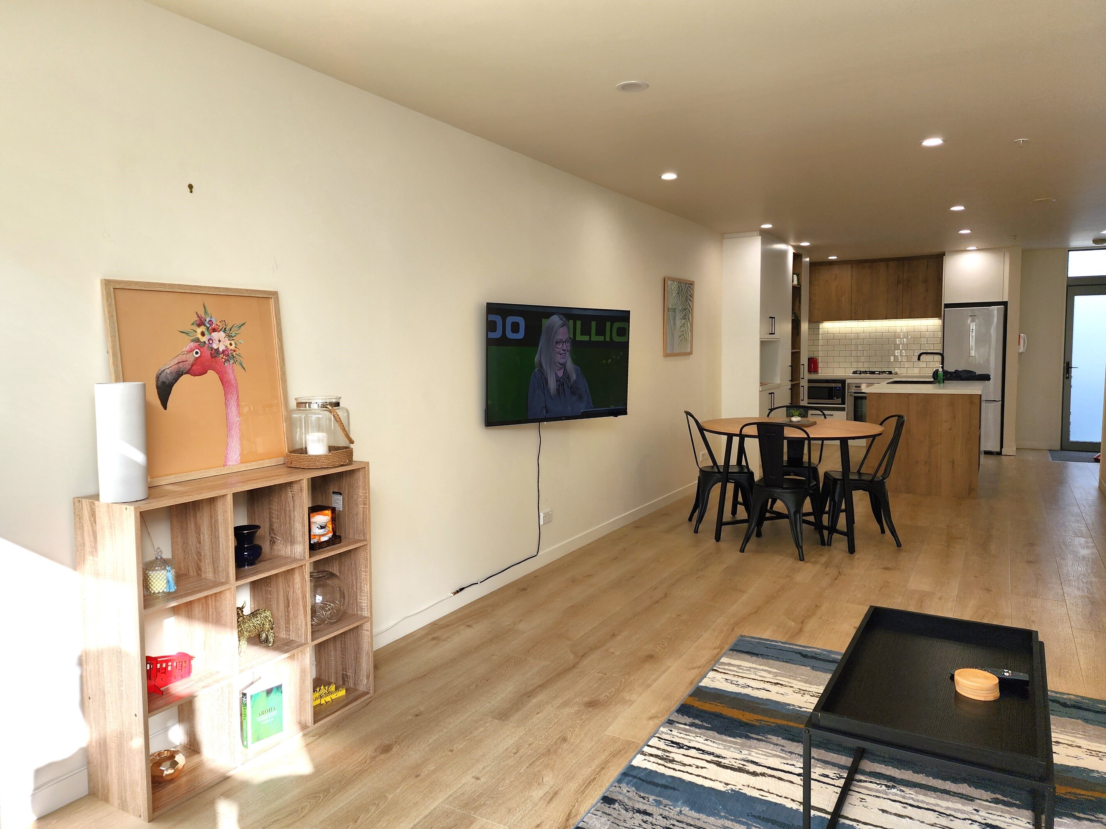 Un salon moderne avec une table à manger en bois, un téléviseur fixé au mur et un coin cuisine à l’arrière-plan.