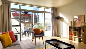 A modern living room with a sofa, coffee table, and a bookshelf. There is a balcony with a view of a building and a fence. - U Suites on Jessie (Wellington)