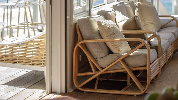 A wicker sofa with cushions near a glass door, overlooking a wooden deck and a view of the ocean.