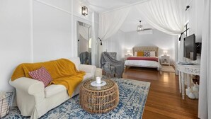 Une chambre d’hôtel moderne avec un canapé blanc, une petite table basse ronde en osier, un lit recouvert d’une courtepointe rouge et blanche, une télévision et une petite table d’appoint.