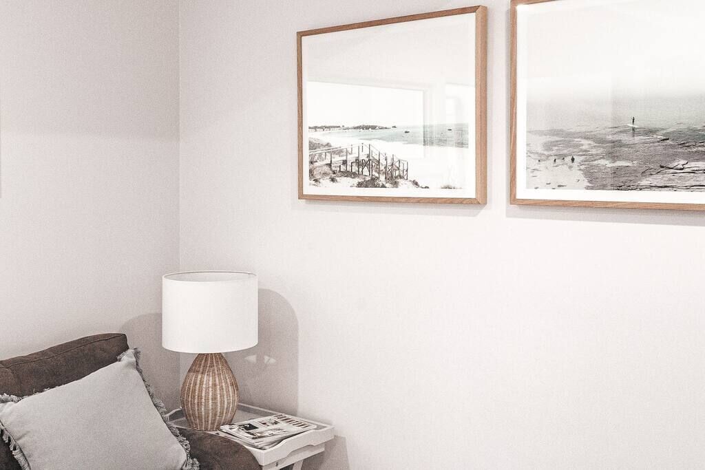 Een kamer met een witte muur, een bruine bank met een grijs kussen, een houten lamp en twee lijstjes met afbeeldingen van kustlandschappen.