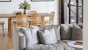 Ein modernes Wohnzimmer mit grauem Sofa, Esstisch aus Holz und Stühlen. An der Wand befinden sich gerahmte Bilder und ein großes Fenster mit Blick.
