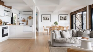 Eine moderne Küche mit weißen Schränken, ein Essbereich mit Holztisch und -stühlen sowie ein graues Sofa mit rundem Couchtisch.