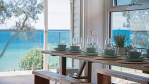 Spisebord dækket med tallerkener, skåle og glas, med udsigt til et naturskønt hav- og trælandskab.