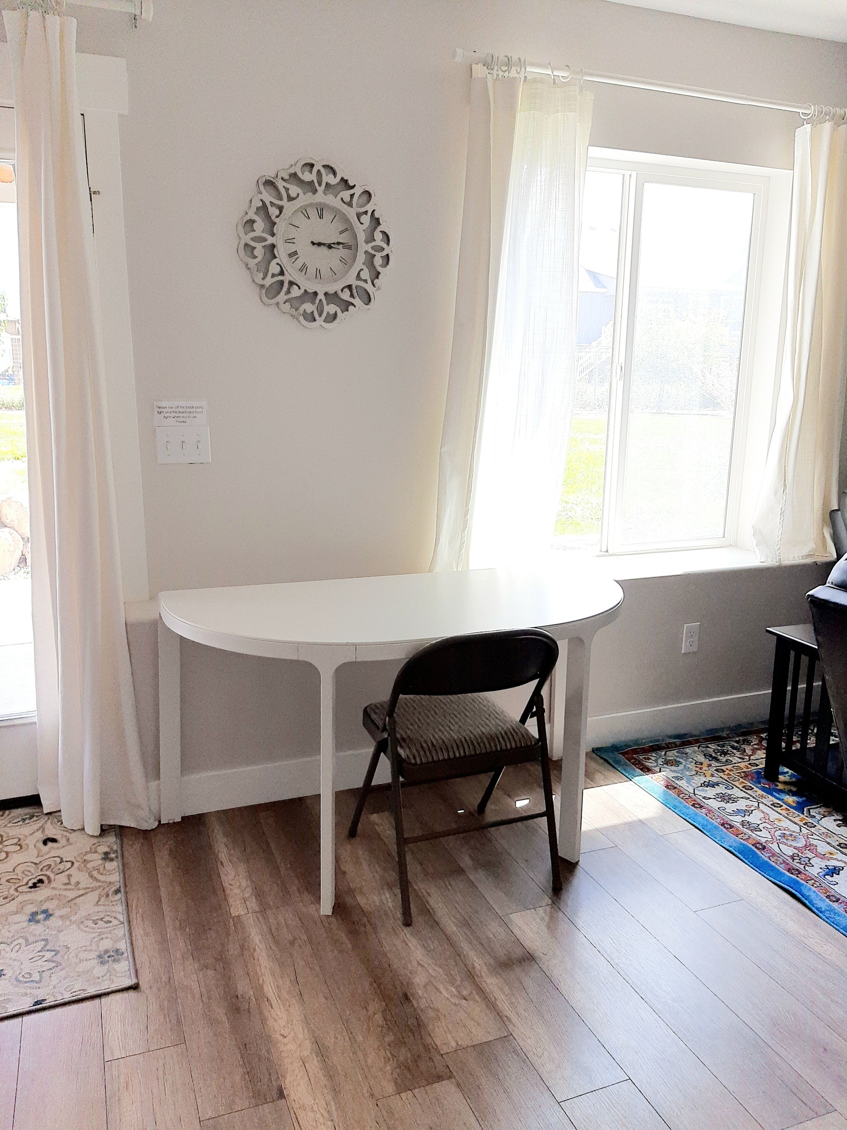 Una habitación con una mesa blanca, una silla negra, un reloj decorativo en la pared y un ventanal con cortinas.