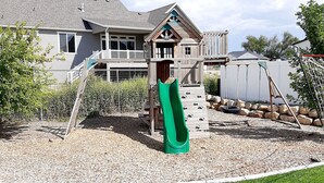 A residential backyard with a wooden play structure, including a slide, and a house in the background.