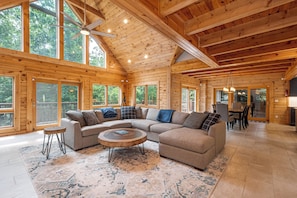 A spacious wooden cabin interior with a large L-shaped sectional sofa, a round coffee table, and a dining area with chairs. - Loch Nest by Taylor Made Deep Creek (McHenry)