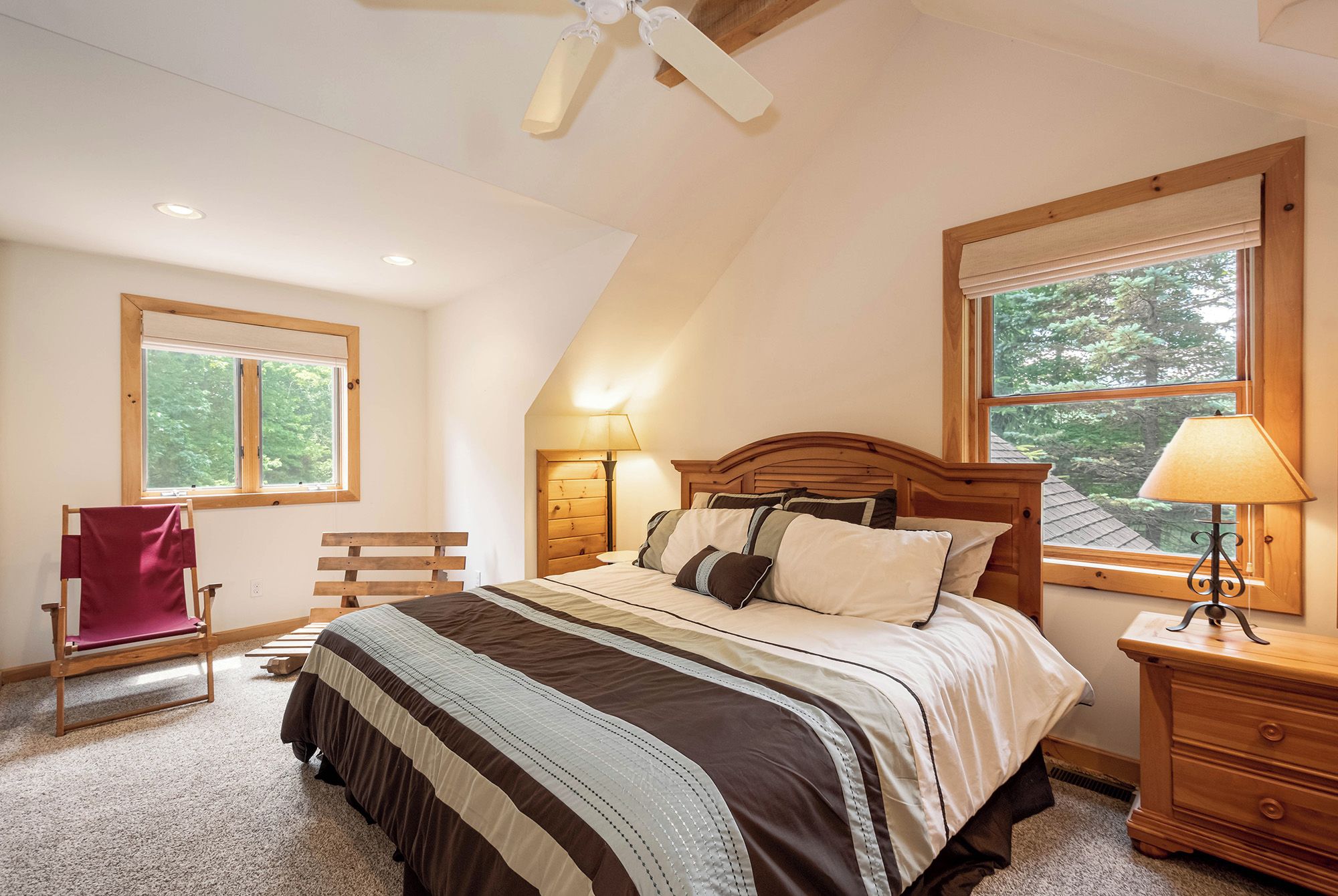 A bedroom with a sloped ceiling, a bed with a wooden headboard, two bedside lamps, a wooden nightstand, a chair, and a window with a view of trees.